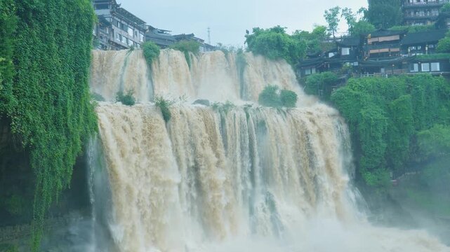 Static wide shot of the massive multi-tiered Furong waterfall rushing beneath ancient town stilt houses