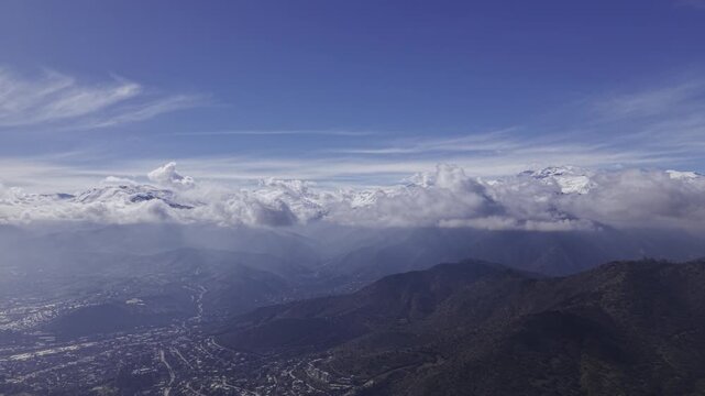 Slow aerial glide above Santiago's eastern edge, framing green foothills, urban neighborhoods and snow-covered Andes behind broken low clouds. Chile, 4K