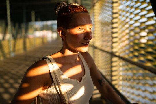 A white woman stands in sunlight and shadow on an urban bridge