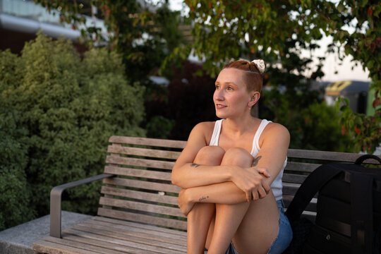 A white woman sits relaxed on a wooden bench in an urban park