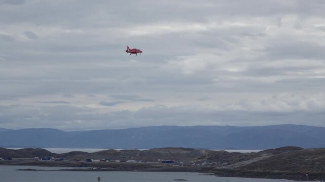 Wide view of an RAF Red Arrows BAE Hawk fast jet on final landing approach, descending through overcast skies over the rocky tundra terrain toward Iqaluit Airport.