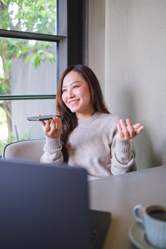 Vertical of a woman holding smart phone and using voice messaging, talking with ai assistant for Artificial intelligence concept while working on laptop