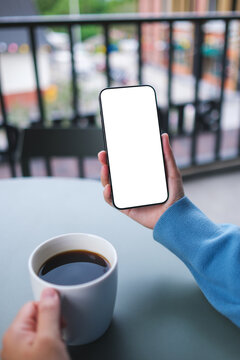 Vertical mockup image of a woman holding and using mobile phone with blank desktop screen while drinking coffee in cafe