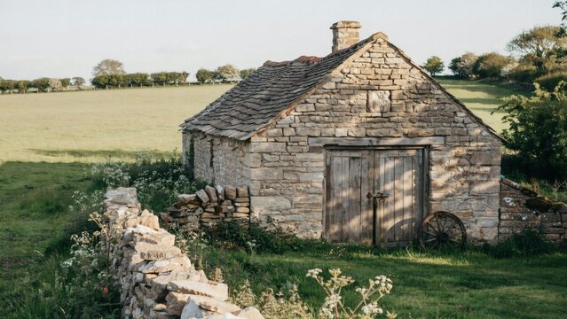 Old stone barn with wooden doors and rustic wheel in a green field with weathered stone building building tiled roof