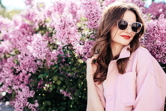 Young beautiful smiling woman in trendy summer clothes. Sexy carefree model posing in the street. Positive model near blooming flower lilac bush beaming. Summertime, pink colours. Red lips, sunglasses