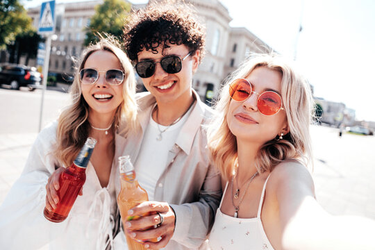 Man and two young beautiful smiling hipster female. Carefree women posing outdoors. Positive models holding and drinking fresh cocktail drink in glass bottle. In the street. Take selfie photos