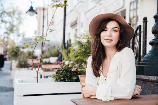 Portrait of beautiful young woman in hat. Model at coffee shop. Female outdoors in sunny day. In white blouse. Cheerful and happy. Waiting for an order at terrace cafe, looks great