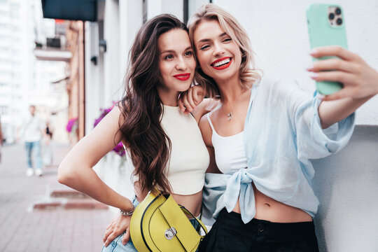 Two young beautiful smiling female in trendy summer clothes. Sexy carefree women posing in street. Positive models having fun, hugging in sunny day. With bright makeup and red lips, take selfie photos