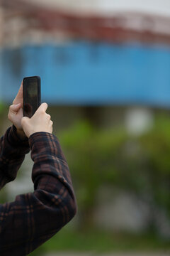 Man photographing blue bridge from street with smartphone held overhead, industrial steel structure and river backdrop, vertical composition, casual