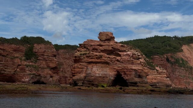 Boat POV gliding past flat topped red sandstone sea stack with balancing capstone standing isolated in tidal waters