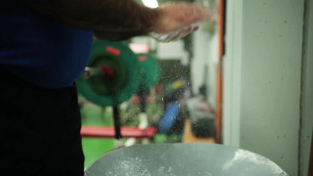 Handheld slow motion close-up, male weightlifter rubbing chalk powder into his hands to improve grip before strength training session, capturing preparation and focus in a gym