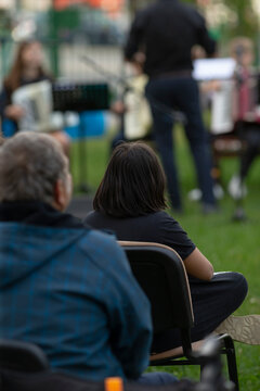 Child and adult watching outdoor concert from lawn chairs, blurred stage and musician in distance, quiet attentive family moment and shared listening