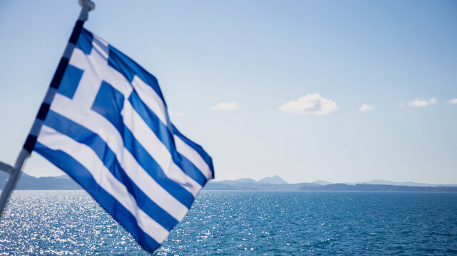 Greek flag billowing from ship above sparkling Aegean Sea water, blue sky, Greece