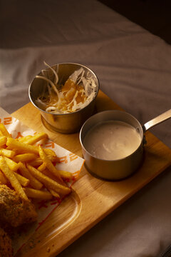 Crispy fries with creamy sauce on wooden board, food on plates styling with shredded cabbage slaw in bowl, breaded fillet bite, metal saucepan of extra dip, warm moody
