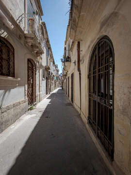 Narrow residential street with historic stone buildings in Ortigia, Syracuse, Sicily, Italy. Old town alleyway with traditional flagstone pavement under a blue sky.
