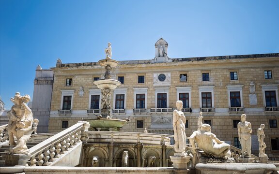 View of the monumental Fontana Pretoria under a bright blue sky, showcasing its multi-tiered marble basins, intricate classical statues, and Palazzo Pretorio in Palermo.