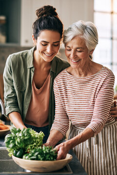 An adult child and their elderly parent standing side-by-side in a bright, modern, accessible kitchen, washing fresh vegetables together in the sink.