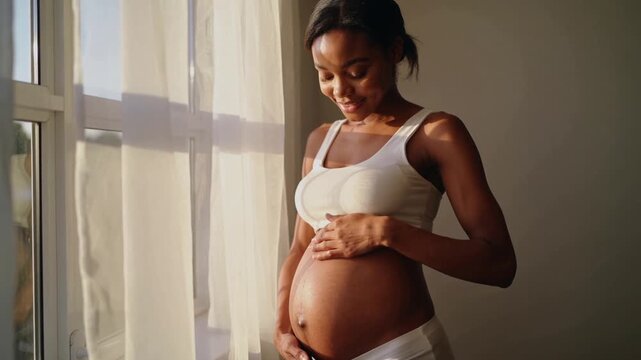 Sunlight drawing pregnant woman in bra, cupping and stroking belly by window drape for bonding