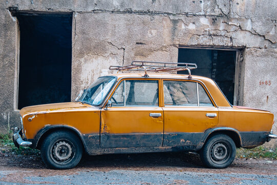 Weathered orange VAZ-2101 Zhiguli, also known as Lada 1200, parked beside a decaying building in Chiatura, Georgia.