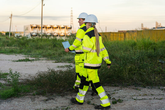 Oil refinery engineers with laptop walking outside industrial plant, petroleum plant workers in safety gear walking together outdoor, energy industry professionals inspecting oil and gas facility site