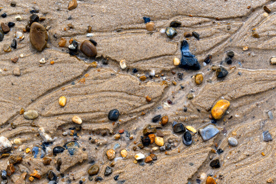 Colourful flint pebbles scattered across wet sand with delicate tidal water patterns at Waxham Beach on the North Sea coast of Norfolk, England.