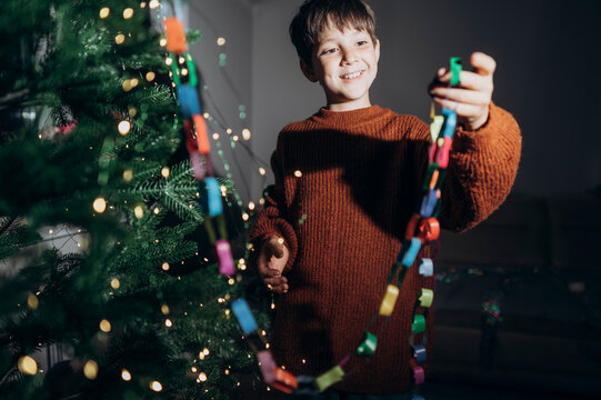 A smiling boy decorates a Christmas tree with a paper chain at home