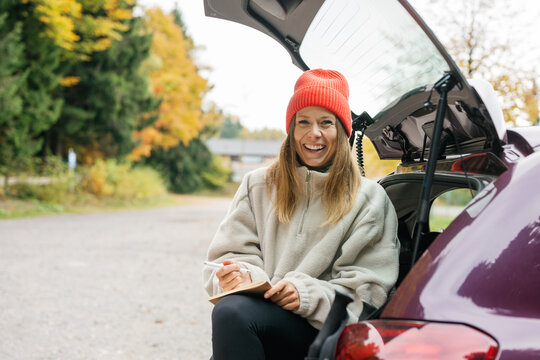 Smiling woman writes in a notebook while sitting in a car trunk