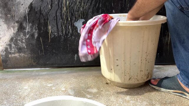 Adult man crouching outdoors washing clothes by hand in a plastic bucket