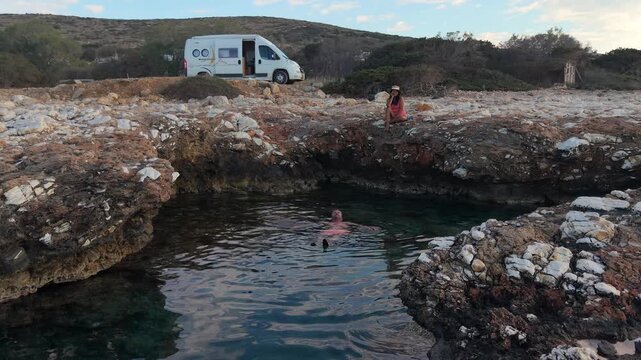 Relaxation comes alive in Paros as people enjoy the crystal-clear waters amidst rocky surroundings. A couple relaxes by the ocean with a camper van motorhome in the background
