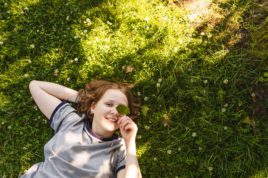 Smiling girl lies in green grass holding a gingko leaf over her eye