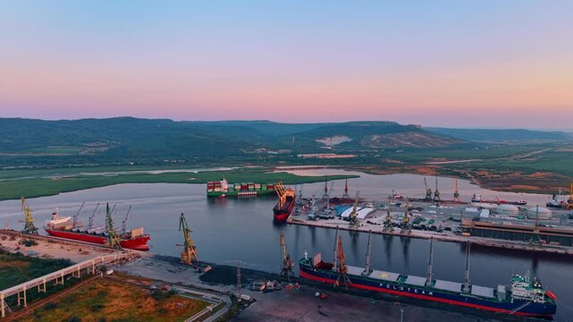 Cargo port with ships and cranes. Large vessel ships sit anchored near dock loading cranes along a river channel under a pink sunset sky.