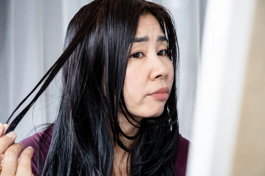 Asian woman worried about oily hair and greasy scalp looking in the mirror. Ideal for hair care products, clarifying shampoo, oil control treatment, scalp hygiene, and beauty