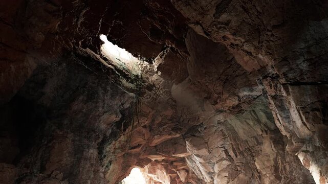 A cavern shows rugged rock walls with a hole at the top. Light filters through this opening, illuminating parts of the cave. It is a natural formation in an outdoor area.