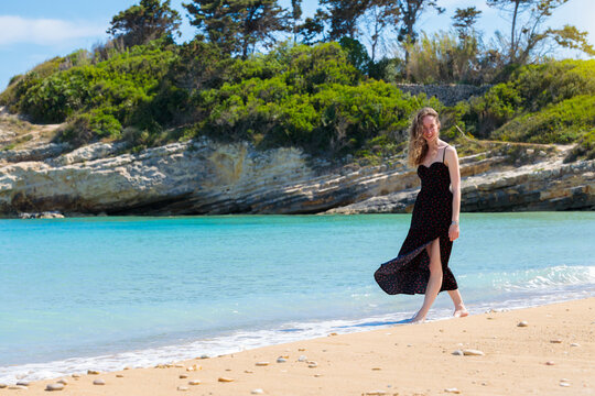 woman walks along the shoreline of a clear turquoise sea, with lush green hills in the background during a summer holiday