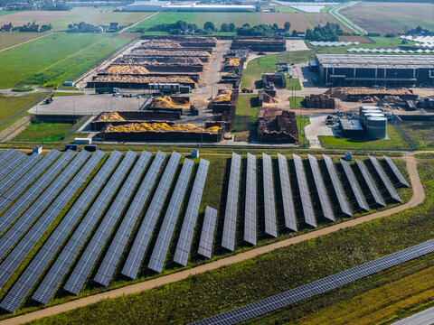 Aerial view of a large solar farm and an adjacent industrial timber processing facility with piles of wood chips and logs in Suwalki, Podlaskie Voivodeship, Poland.
