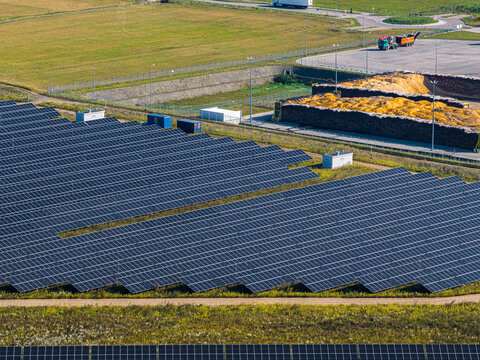 Aerial view of a large solar farm with rows of photovoltaic panels installed in a grassy field near an industrial site with wood piles in Suwalki, Podlaskie Voivodeship, Poland.