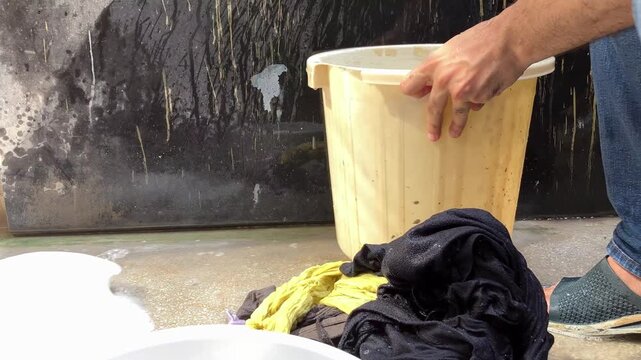 Adult man crouching outdoors washing clothes by hand in a plastic bucket