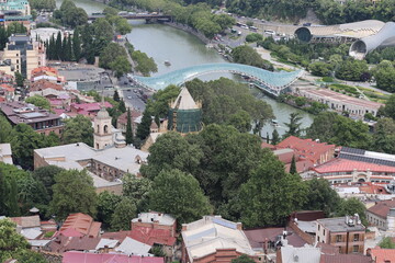Aerial view of Tbilisi, Georgia with Peace Bridge over Kura river, old town rooftops, church towers and Rike park. Cityscape with lush greenery. © Ольга Дербасова