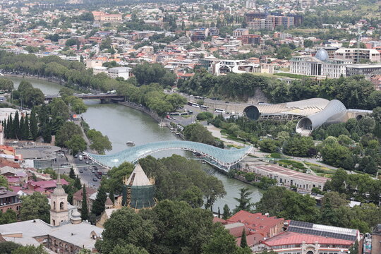 Aerial view of Tbilisi, Georgia with Peace Bridge over Kura river, old town rooftops, church towers and Rike park among lush greenery.