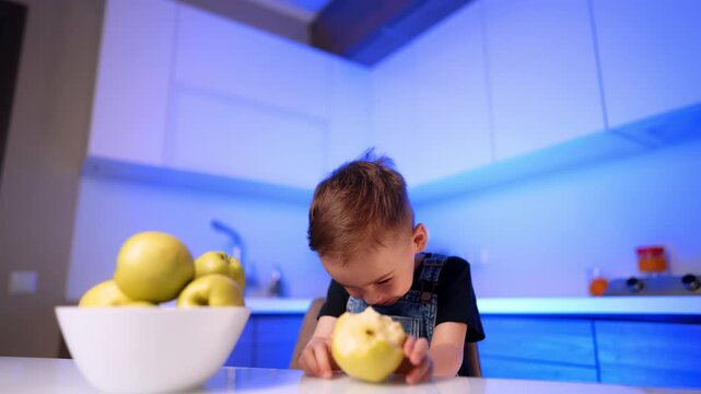 Boy leaning forward to eat yellow fruit at table. A child wearing denim overalls bites an apple near a bowl in a blue lit kitchen environment.