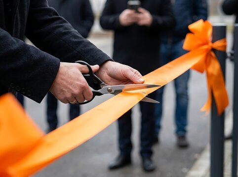 Man in black coat cutting orange ribbon with scissors for grand opening event with blurred people in background celebrating achievement.