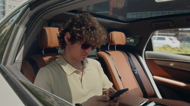 Caucasian young adult man sitting in backseat of moving car, using smartphone. Tracking shot perfect for ride-sharing app or business travel promotions