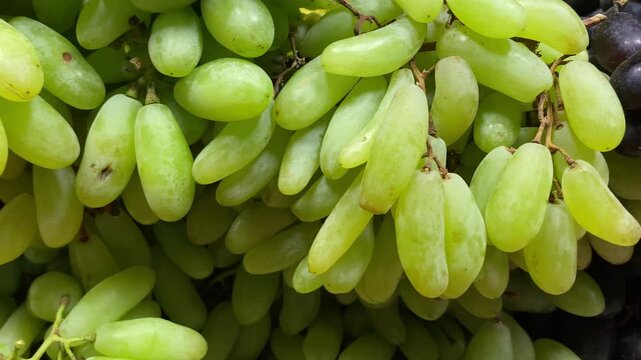 Macro close-up shot of fresh green elongated seedless grapes bundled together, highlighting texture, vibrant color, and fresh produce displayed at a local market stand in bright daylight.
