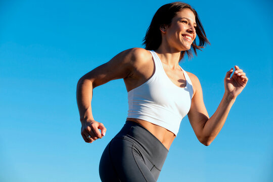 Young adult Caucasian woman jogging outdoors against clear blue sky, promoting active lifestyle. Image suitable for fitness campaigns and wellness marketing, low angle shot