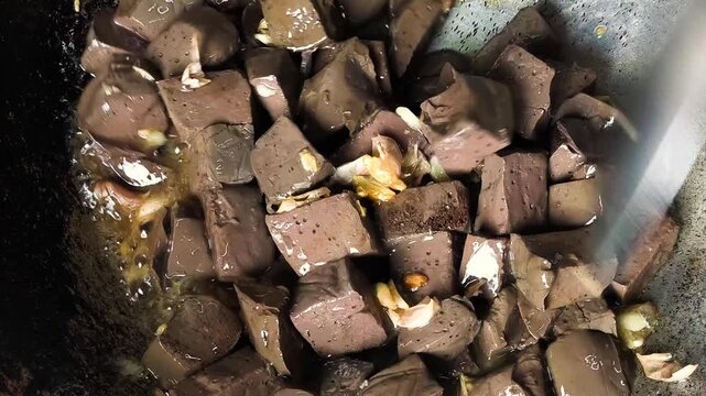 Dynamic high-angle shot of a chef cooking cubed chicken blood curd in a traditional wok. The brown, porous blocks are being flipped and saut&eacute;ed in oil along with aromatic crushed garlic. This footage 