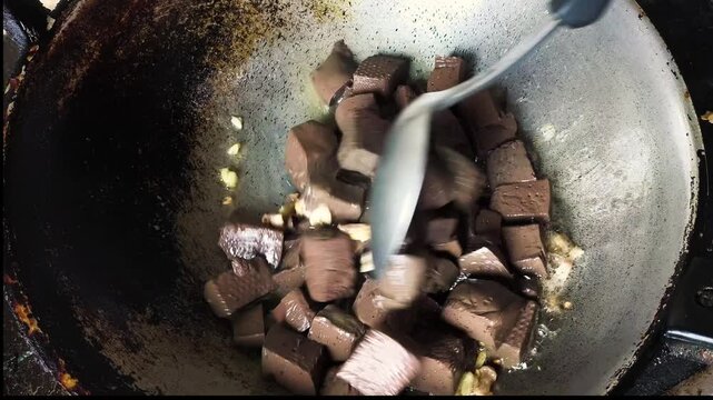 Dynamic high-angle shot of a chef cooking cubed chicken blood curd in a traditional wok. The brown, porous blocks are being flipped and saut&eacute;ed in oil along with aromatic crushed garlic. This footage 