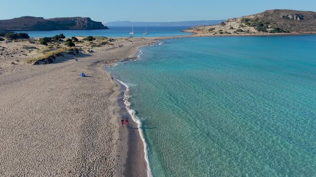 Discover the stunning landscape of Simos Beach in Elafonisos Greece as visitors enjoy sunbathing and walking along the crystal-clear waters under a bright blue sky.