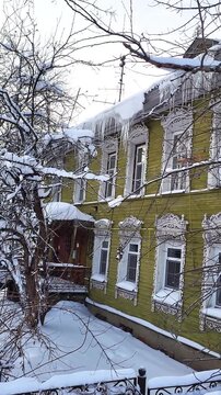 An old wooden house stands surrounded by deep snowdrifts during a cold winter season. Long icicles are hanging from the edge of the roof, and the ground is covered with a thick layer of snow.