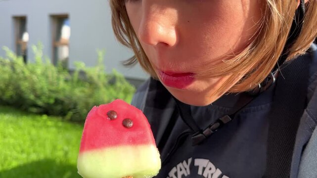 A close up shot of a young boy taking a bite from a watermelon ice lolly outside, food industry, advertising, summer refreshment, delicious frozen snack, outdoor fun.