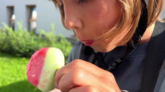 A young boy looking down while eating a red and green ice lolly in the garden, food blog, e-commerce, summer heat, delicious ice cream, casual outdoor moment.